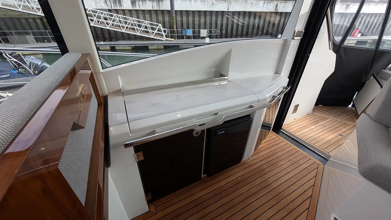 A sleek yacht interior featuring a white countertop, black fridge, wooden flooring, and a view of the marina outside.